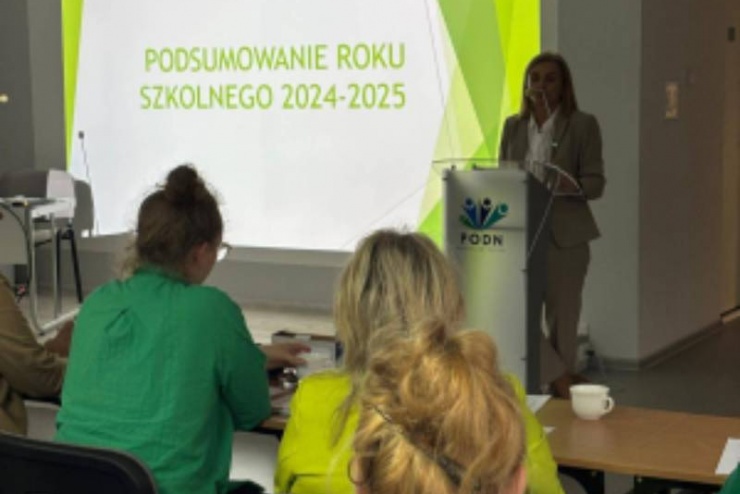 Na zdjęciu pokazana jest kobieta stojąca przy mównicy w tle z rzutnika jest prezentowana prezentacja. Przed mównicą siedzą dwie kobiety.