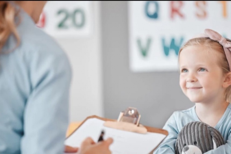 Na zdjęciu pokazana jest Dziewczynka uśmiechająca się w stronę Pani, która trzyma w lewej dłoni dokumenty a w prawej długopis.