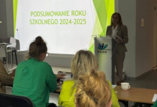 Na zdjęciu pokazana jest kobieta stojąca przy mównicy w tle z rzutnika jest prezentowana prezentacja. Przed mównicą siedzą dwie kobiety.