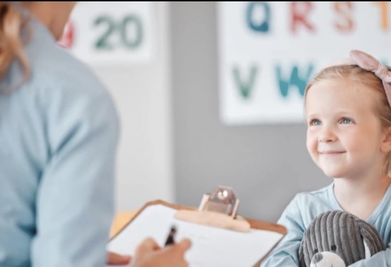Na zdjęciu pokazana jest Dziewczynka uśmiechająca się w stronę Pani, która trzyma w lewej dłoni dokumenty a w prawej długopis.