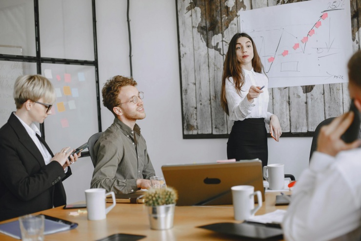 Jest grupa na zdjęciu trzy osoby siedzą przy stole a osoba stojąca do nich przemawia