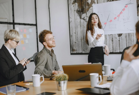 Jest grupa na zdjęciu trzy osoby siedzą przy stole a osoba stojąca do nich przemawia